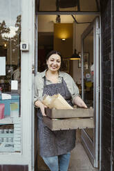 Portrait of happy female owner standing at doorway of deli holding crate - MASF33808