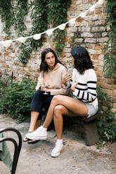 Female friends talking to each other while sitting on bench near brick wall at garden party - MASF33825