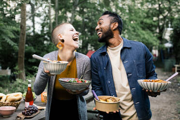 Multiracial male and female friends laughing and looking at each other while holding food bowls - MASF33833