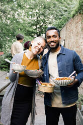 Portrait of smiling male and female friends holding food bowls while standing in back yard - MASF33834
