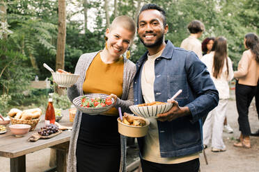 Portrait of smiling friends holding food bowls during dinner party in back yard - MASF33835