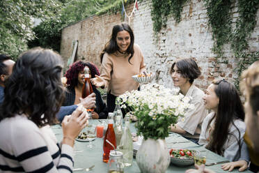 Smiling woman holding bowl while talking to male and female friends during dinner party in back yard - MASF33845