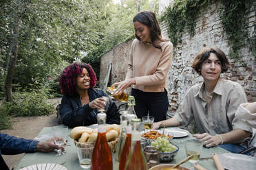 Woman serving drink to female friend sitting at dining table during dinner party in back yard - MASF33847