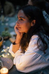 Woman with hand on chin sitting at dining table by illuminated candle - MASF33879