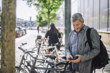 Smiling mature businessman using smart phone while standing at bicycle parking station - MASF33904