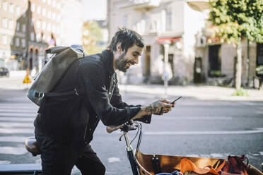 Side view of smiling businessman using smart phone while leaning on cargo bike - MASF33908