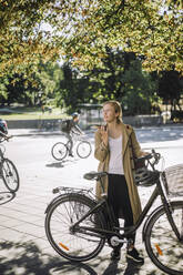 Junge Pendlerin, die mit ihrem Fahrrad unter einem Baum steht und über einen Lautsprecher telefoniert - MASF33911