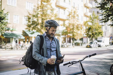 Mature businessman with disposable cup using smart phone by bicycle - MASF33914