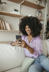 Lächelnde Afro-Frau, die ein Smartphone benutzt und zu Hause auf dem Sofa sitzt - RCPF01589