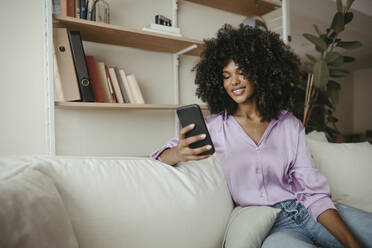 Junge Frau mit Afrofrisur, die im Wohnzimmer ein Mobiltelefon benutzt - RCPF01590