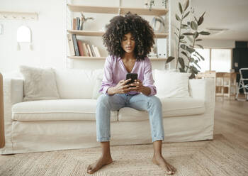 Young woman using mobile phone sitting on sofa in living room - RCPF01591