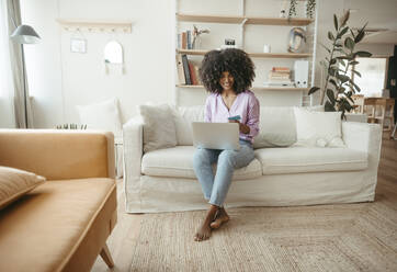 Smiling woman paying with credit card on laptop in living room - RCPF01598