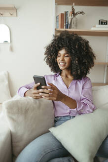 Smiling woman using smart phone sitting on sofa at home - RCPF01599