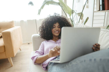 Smiling Afro woman using laptop lying on sofa at home - RCPF01612