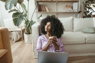 Thoughtful woman with coffee cup and laptop in living room - RCPF01615