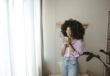 Smiling woman holding coffee cup day dreaming at home - RCPF01616