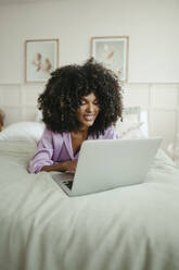 Smiling woman using laptop lying on bed in bedroom - RCPF01621