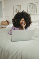Junge Frau mit Hand am Kinn, die einen Laptop im Schlafzimmer benutzt - RCPF01623