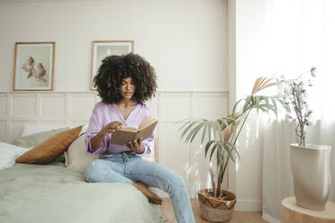 Afro woman reading book sitting on bed in bedroom - RCPF01628