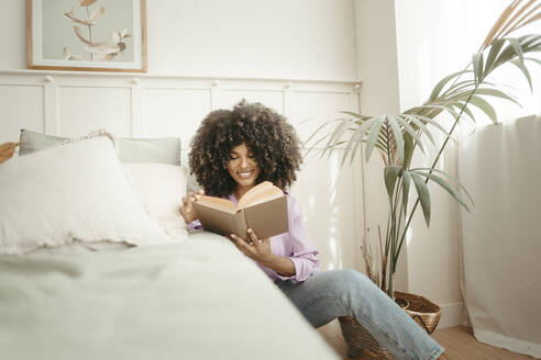 Lächelnde Frau, die ein Buch liest, sitzt am Bett im Schlafzimmer - RCPF01630