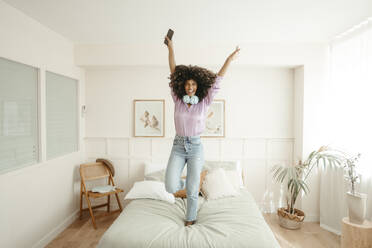 Happy woman with arms raised dancing on bed in bedroom - RCPF01632