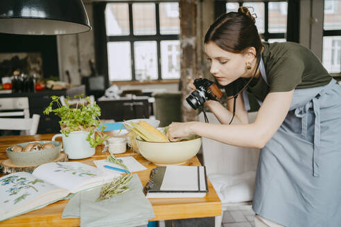 Food-Stylistin mit Digitalkamera, die eine Gemüseschale auf einem Tisch im Studio aufstellt - MASF33930