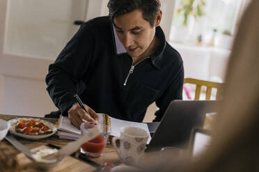 Young male freelancer taking down notes while talking on smart phone at dining table - MASF33939