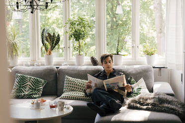 Junger Mann liest Zeitung, während er zu Hause auf dem Sofa im Wohnzimmer sitzt - MASF33948