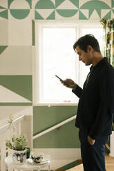 Seitenansicht eines Mannes mit Hand in der Tasche, der sein Smartphone benutzt, während er zu Hause steht - MASF33956