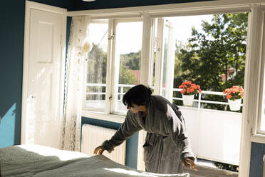 Frau im Bademantel, die ein Bettlaken auf das Bett legt und sich gegen die Balkontür zu Hause beugt - MASF33960