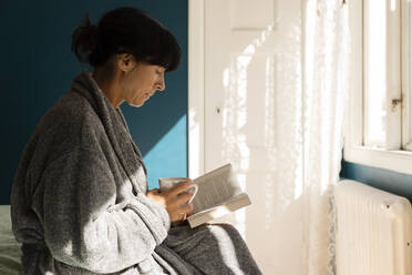 Frau im Bademantel, die eine Tasse hält, während sie im Schlafzimmer zu Hause ein Buch liest - MASF33969