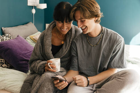 Glückliche Mutter und Sohn teilen sich ein Smartphone, während sie auf dem Bett im Schlafzimmer sitzen - MASF33970