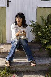 Happy woman holding cup looking away while sitting on porch outside house - MASF33984