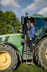 Glücklicher reifer Landwirt steht auf einem Traktor auf einem Bauernhof unter bewölktem Himmel - MASF34029