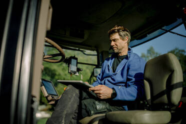 Älterer Landwirt mit Tablet-PC im Traktor sitzend an einem sonnigen Tag - MASF34031