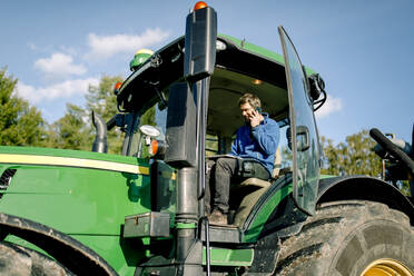 Landwirt benutzt einen Tablet-PC und spricht mit seinem Smartphone, während er im Traktor auf dem Bauernhof sitzt - MASF34032