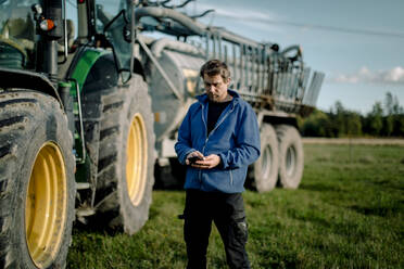 Älterer Landwirt, der ein Smartphone benutzt und auf einem Feld vor einem Traktor steht - MASF34036