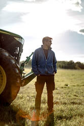 Landwirt mit den Händen in den Taschen, der an einem sonnigen Tag am Traktor steht - MASF34037