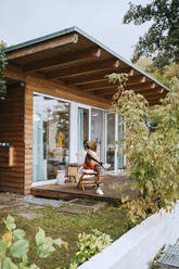 Smiling mature man sitting on chair on porch outside tiny house - MASF34093