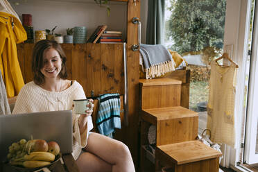 Lächelnde junge Frau mit Laptop und Kaffeetasse, während sie zu Hause sitzt - MASF34099