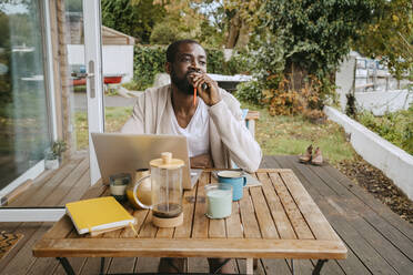 Nachdenklicher Mann mit Laptop sitzt am Tisch auf der Veranda - MASF34107
