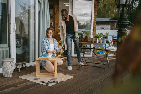 Älterer Mann betrachtet Frau, die Möbel auf der Veranda vor dem Haus streicht - MASF34120
