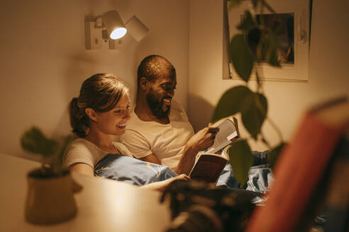 Lächelndes Paar, das bei einer beleuchteten Wandlampe zu Hause Bücher liest - MASF34135