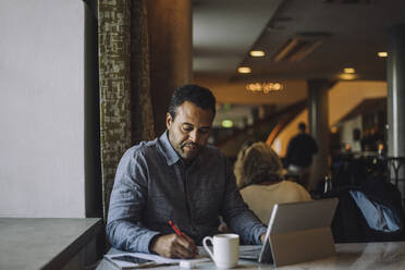 Male freelancer writing in diary while sitting with laptop at table - MASF34143