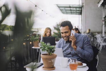 Smiling male entrepreneur talking on smart phone while sitting with laptop at cafe - MASF34147