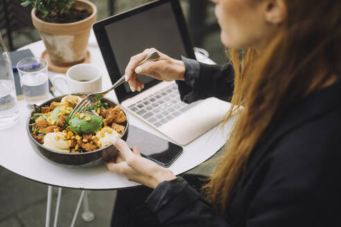Geschäftsfrau mit Schüssel mit Essen und Laptop am Tisch - MASF34150