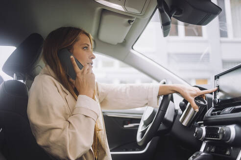 Weibliche Unternehmerin, die über ihr Mobiltelefon spricht, während sie das Touchscreen-Bedienfeld auf dem Armaturenbrett im Auto benutzt - MASF34153