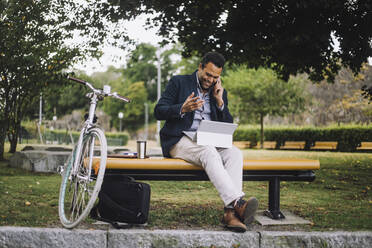 Männlicher Freiberufler in voller Länge, der im Park sitzend mit seinem Smartphone spricht - MASF34164