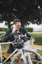 Happy businessman talking on mobile phone while sitting by bicycle at park - MASF34168