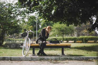 Geschäftsfrau in voller Länge, die mit ihrem Smartphone telefoniert, während sie im Park am Fahrrad sitzt - MASF34169